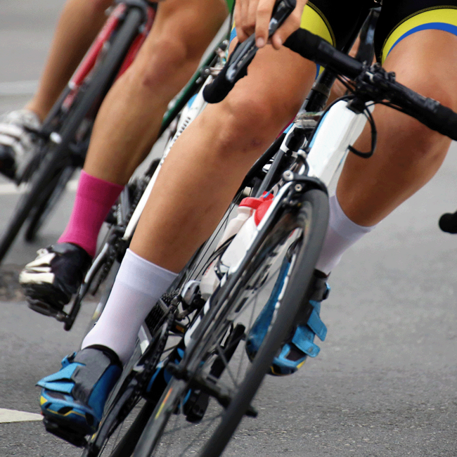 Drei Rennradfahrer die hintereinander fahren. Man sieht aufgrund des Bildausschnitts nur die Fahrräder und Beine der Sportler*innen.