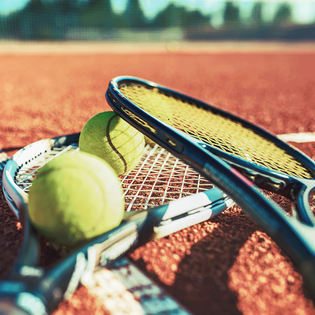 Zwei Tennisschläger und zwei Tennisbälle liegen neben einem im Hintergrund unfokussiert zu sehendem Tennisfeld.