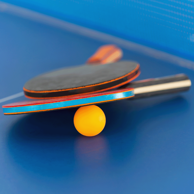 Zwei Tischtennisschläger liegen auf einem orangenem Tischtennisball. Diese liegen alle auf einer blauen Oberfläche