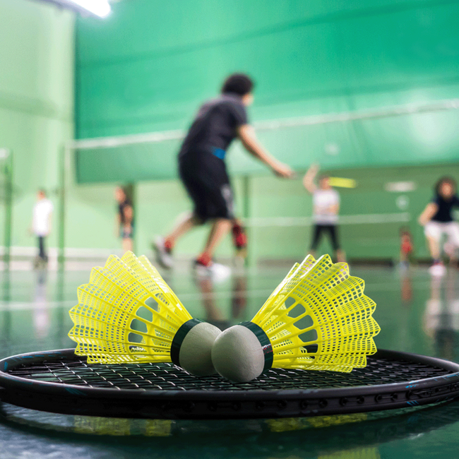Ein Badmintonschläger, der auf dem Boden einer Turnhalle liegt und auf dem zwei Badmintonbälle liegen. Im Hintergrund sieht man einige Personen die Badminton trainieren.
