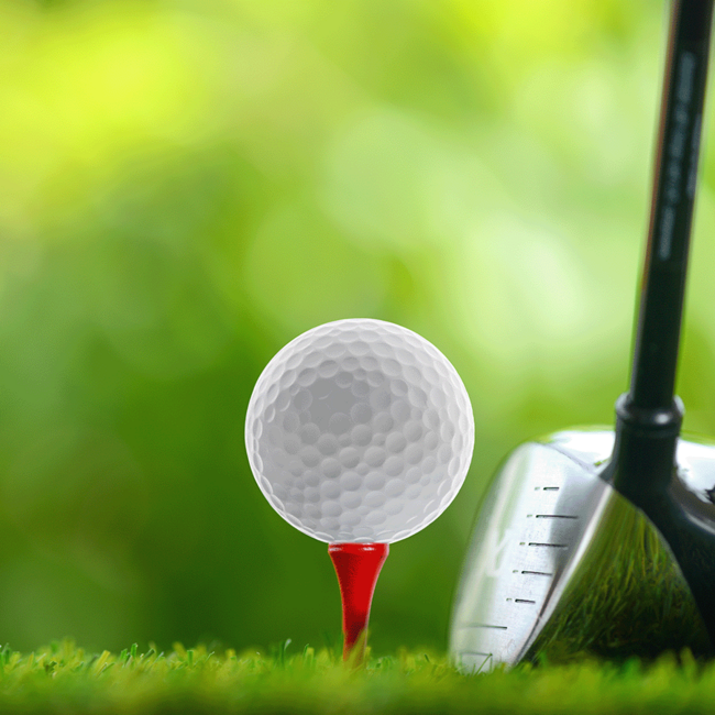 Ein Golfball mit dem Kopf eines Golfschlägers rechts daneben vor einem verschwommenen grünen Hintergrund.